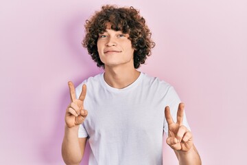 Handsome young man wearing casual white t shirt smiling looking to the camera showing fingers doing...