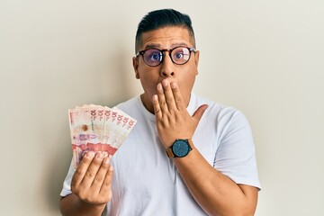 Young latin man holding 10 colombian pesos banknotes covering mouth with hand, shocked and afraid...