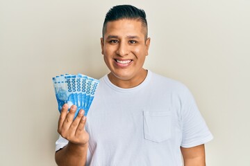 Young latin man holding 10000 chilean pesos looking positive and happy standing and smiling with a confident smile showing teeth