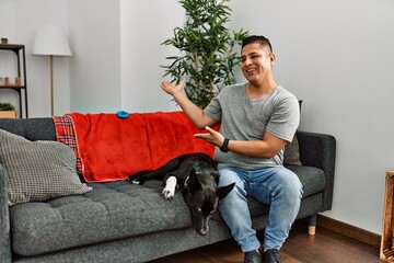 Young latin man and dog sitting on the sofa at home inviting to enter smiling natural with open hand
