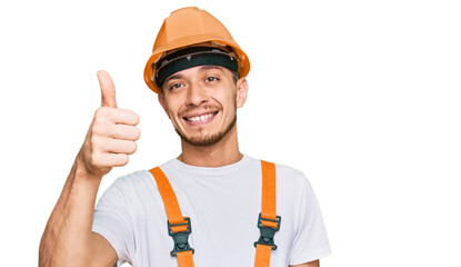 Hispanic young man wearing handyman uniform and safety hardhat smiling happy and positive, thumb up doing excellent and approval sign
