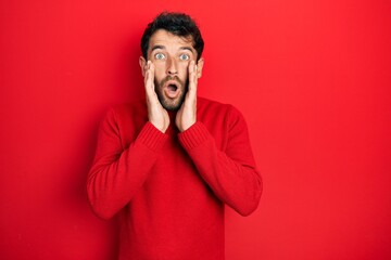 Handsome man with beard wearing casual red sweater afraid and shocked, surprise and amazed expression with hands on face