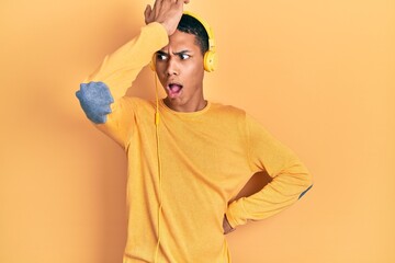 Young african american guy listening to music using headphones surprised with hand on head for...