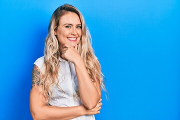 Beautiful young blonde woman wearing casual white shirt looking confident at the camera smiling with crossed arms and hand raised on chin. thinking positive.