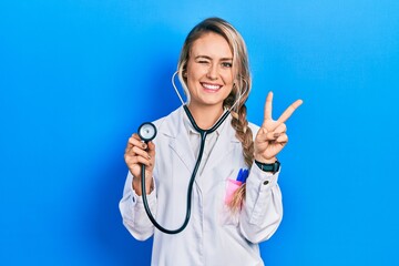 Beautiful young blonde doctor woman holding stethoscope smiling with happy face winking at the...