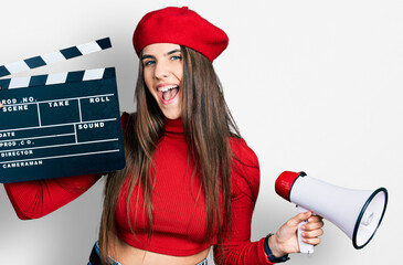 Young brunette teenager holding video film clapboard and megaphone smiling and laughing hard out...
