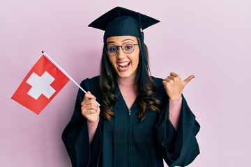 Young hispanic woman wearing graduation uniform holding switzerland flag pointing thumb up to the side smiling happy with open mouth