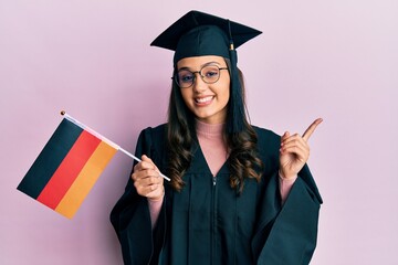 Young hispanic woman wearing graduation uniform holding germany flag smiling happy pointing with hand and finger to the side