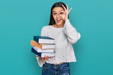 Young hispanic girl holding a pile of books smiling happy doing ok sign with hand on eye looking through fingers