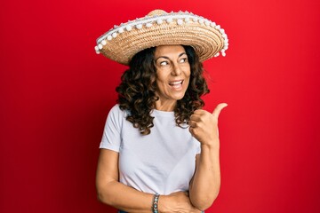 Middle age hispanic woman holding mexican hat smiling with happy face looking and pointing to the side with thumb up.