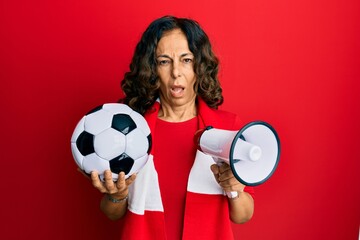 Middle age hispanic woman football hooligan holding ball and using megaphone clueless and confused...