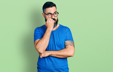 Hispanic man with beard wearing casual t shirt and glasses looking stressed and nervous with hands on mouth biting nails. anxiety problem.