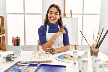 Young brunette woman at art studio with painted hands with a big smile on face, pointing with hand and finger to the side looking at the camera.
