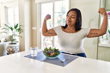 Young hispanic woman eating healthy salad at home showing arms muscles smiling proud. fitness concept.