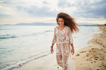 Portrait of smiling young curly woman on white dress walking on beach