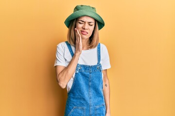 Young caucasian blonde woman wearing denim jumpsuit and hat with 90s style touching mouth with hand...