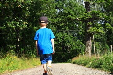 Kid with blue shirt walking outside along a graveled road. Back towards camera. Green nature and grass on the sides. Stockholm, Sweden, Europe