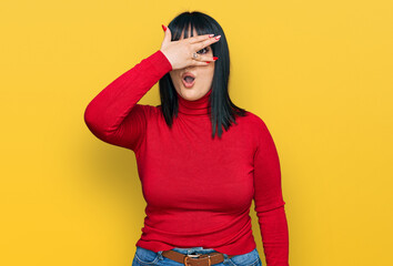 Fototapeta premium Young hispanic woman wearing casual clothes peeking in shock covering face and eyes with hand, looking through fingers with embarrassed expression.