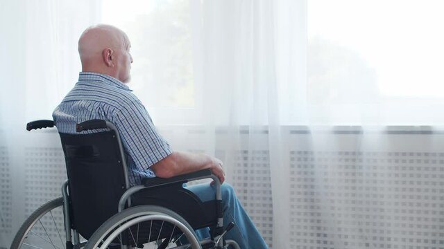 Disabled old man is sitting in a wheelchair at home alone. A handicapped person in a wheelchair is looking out the window. Paralysis and disability.