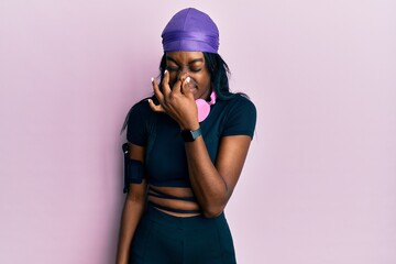 Young african american woman wearing gym clothes and using headphones smelling something stinky and disgusting, intolerable smell, holding breath with fingers on nose. bad smell