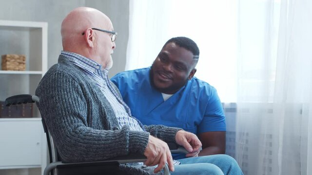 African-American caregiver and old disabled man in a wheelchair. Professional nurse and handicapped patient in a nursing home. Assistance, rehabilitation and health care.