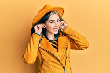 Young modern girl wearing yellow hat and leather jacket smiling pulling ears with fingers, funny...