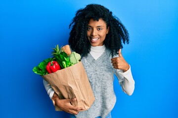 African american woman with afro hair holding paper bag with groceries smiling happy and positive, thumb up doing excellent and approval sign