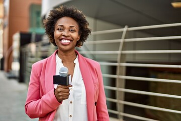 Young african american woman journalist holding reporter microphone speaking and smiling to the...