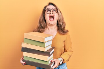 Middle age caucasian woman holding a pile of books angry and mad screaming frustrated and furious, shouting with anger looking up.