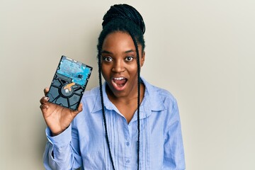 African american woman with braided hair holding hard disk scared and amazed with open mouth for...