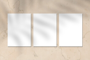 Three empty white vertical rectangle poster or business card mockup with tropical leave shadow on beige marble stone wall. Flat lay, top view. For advertising, brand design, stationery presentation.