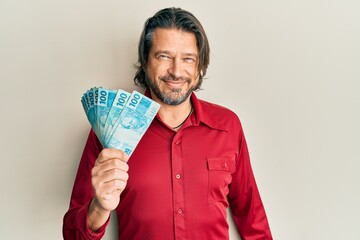 Middle age handsome man holding 100 brazilian real banknotes looking positive and happy standing...