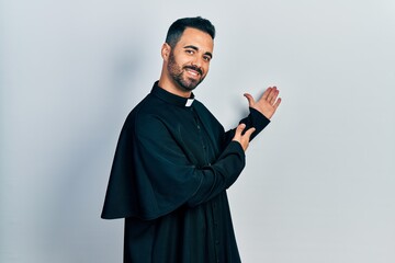 Handsome hispanic man with beard wearing catholic priest robe inviting to enter smiling natural...