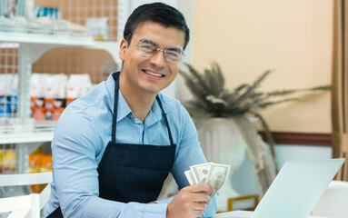 Fototapeta premium Caucasian handsome male cafe business owner or entrepreneur wearing blue apron and eyeglasses, smiling with happiness and success, holding money in his hand, calculating profits while sitting in shop.