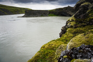 rocky cliff of basalt on the river bank, the banks are covered with green moss, cloudy sky, nature of Iceland