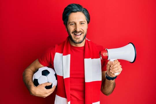 Young hispanic man football hooligan holding ball using megaphone smiling and laughing hard out loud because funny crazy joke.