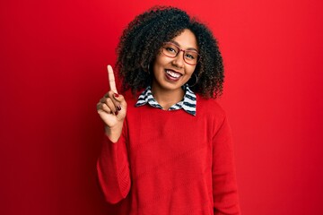 Beautiful african american woman with afro hair wearing sweater and glasses pointing finger up with...