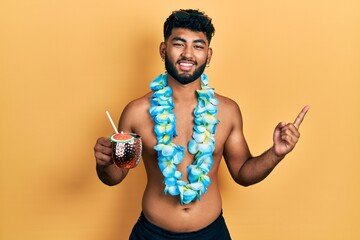 Arab man with beard wearing swimsuit and hawaiian lei drinking cocktail smiling happy pointing with...