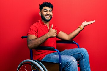 Arab man with beard sitting on wheelchair showing palm hand and doing ok gesture with thumbs up,...
