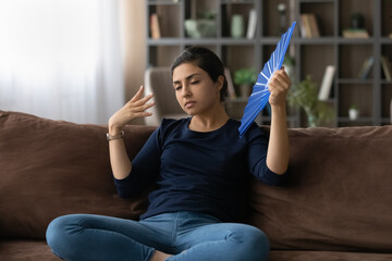 Tired millennial Indian woman sit rest on sofa at home feel unwell suffer from heatstroke wave with...