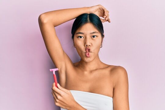 Young chinese woman wearing shower towel using razor making fish face with mouth and squinting eyes, crazy and comical.
