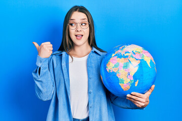 Young hispanic girl holding world ball pointing thumb up to the side smiling happy with open mouth