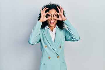 Young latin girl wearing business clothes and glasses doing ok gesture like binoculars sticking tongue out, eyes looking through fingers. crazy expression.