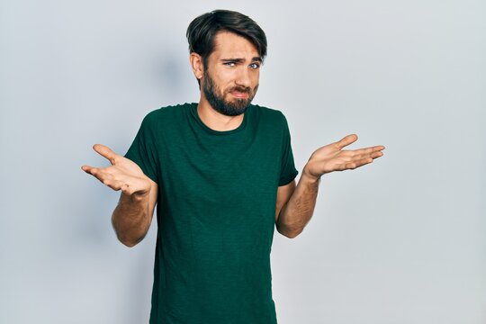Young hispanic man wearing casual white tshirt clueless and confused expression with arms and hands raised. doubt concept.