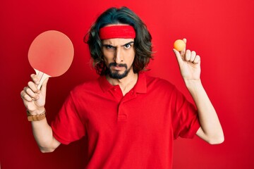 Young hispanic man holding red ping pong racket and ball skeptic and nervous, frowning upset...