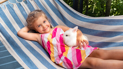 A beautiful girl with positive emotions lies on a hammock and holds a dwarf rabbit in her hands....