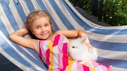 A girl with positive emotions lies on a hammock and holds a white decorative rabbit in her hands. A...