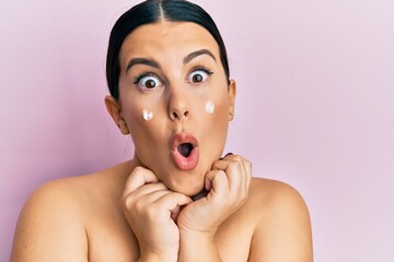 Beautiful brunette woman standing topless wearing face cream afraid and shocked with surprise and...