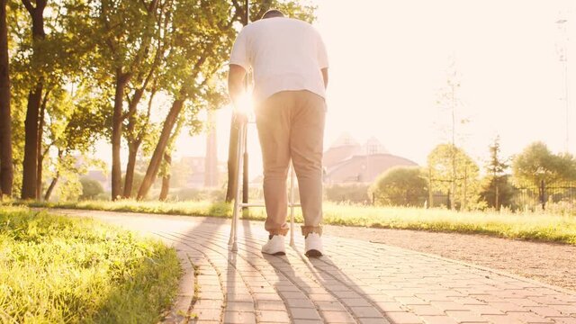 Disabled African-American man is walking with a walker. Handicapped patient in the park. Assistance, rehabilitation and health care. Golden hour sunset.