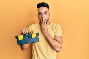 Young arab man holding toolbox covering mouth with hand, shocked and afraid for mistake. surprised...